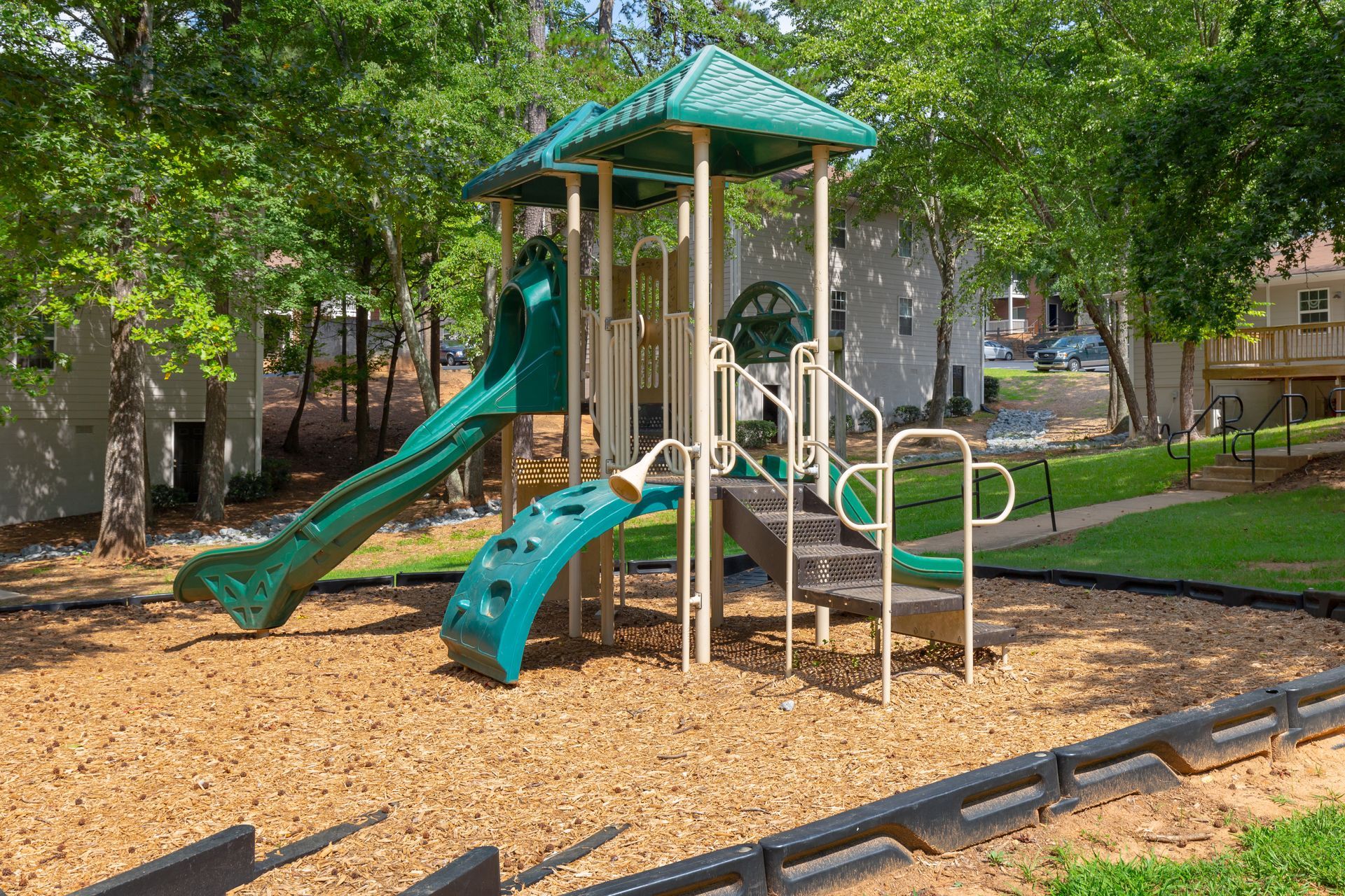 A green and beige modular playground structure with a slide, climbing wall, and stairs on wood chips in a wooded area.