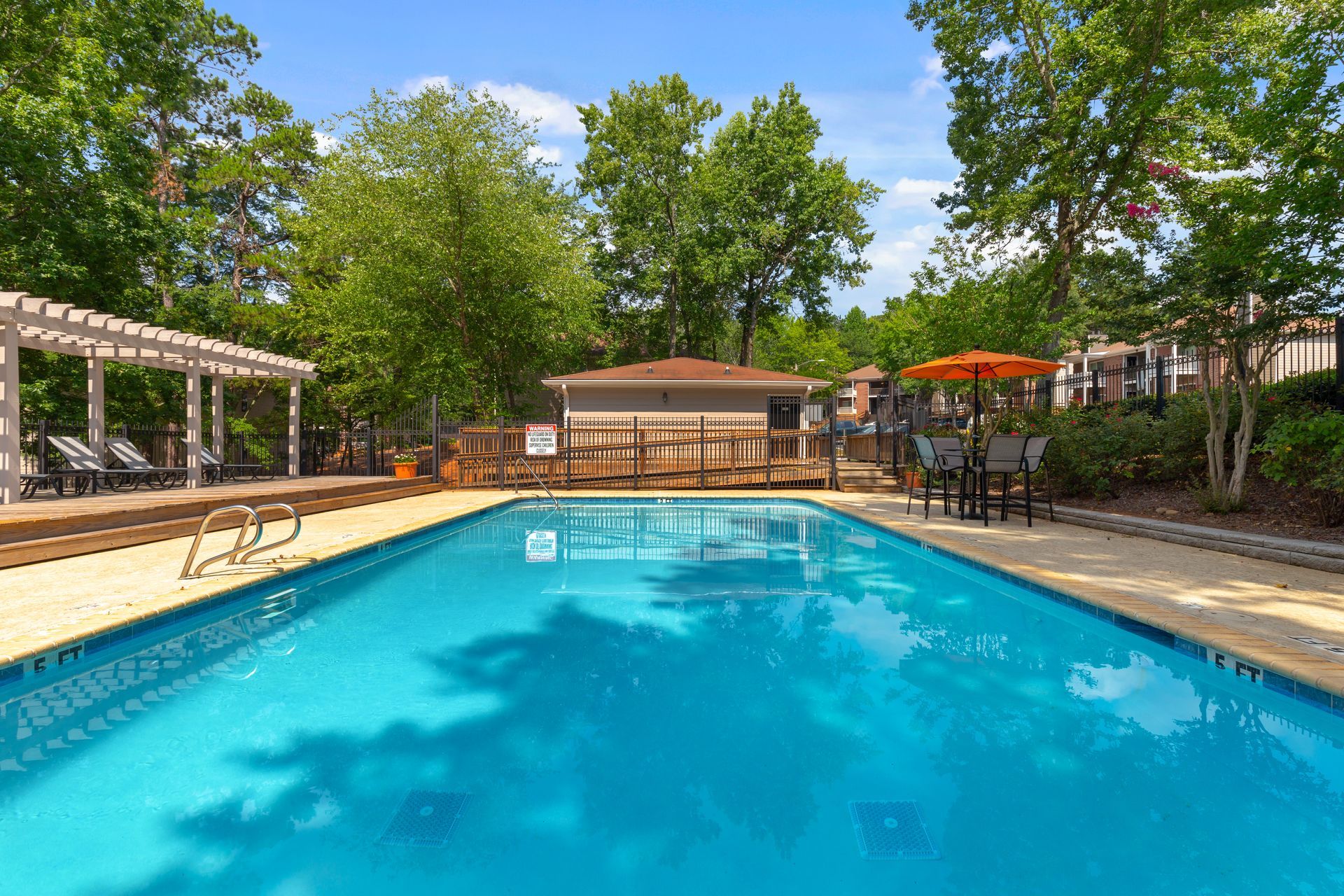 An outdoor swimming pool with blue water, surrounded by trees, a wooden pergola, and a small building under a blue sky.