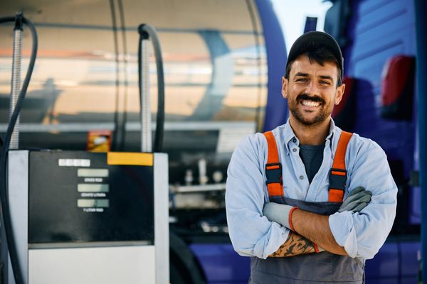 A man is standing in front of a truck at a gas station with his arms crossed.