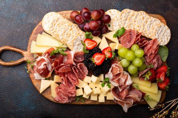 A wooden cutting board topped with meat , cheese , fruit and crackers.