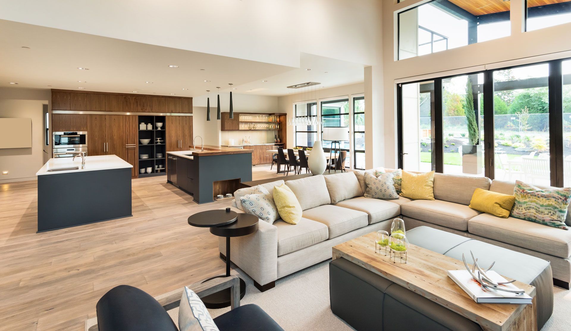 Spacious modern living room with kitchen and large windows; neutral colors, sectional sofa, and wood accents.