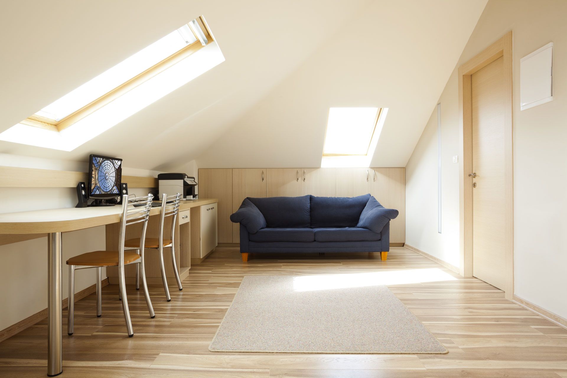 A bright, airy attic room with a desk, blue sofa, rug, and two skylights.