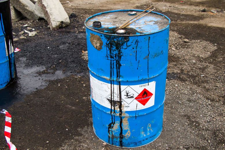 Blue hazardous waste barrel with white label and black spill stains on a dirty ground.