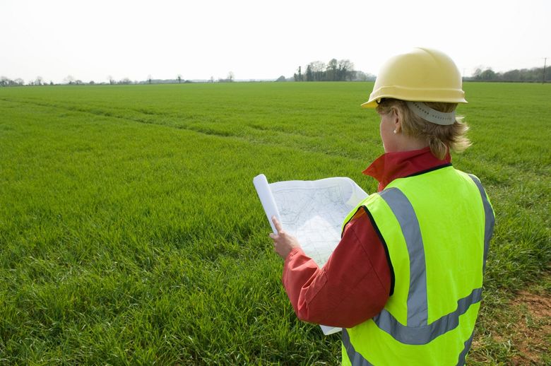 Worker in yellow hard hat and reflective vest inspecting a green field with a clipboard