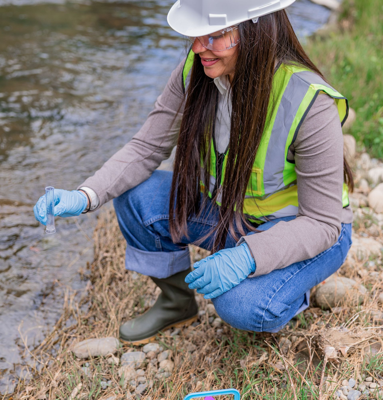 Worker in safety vest and hard hat crouches by a stream, sampling water with blue gloves.