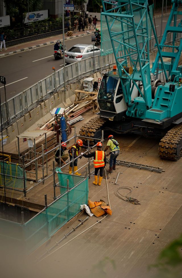 Construction workers beside a turquoise excavator at a roadside worksite behind metal fencing