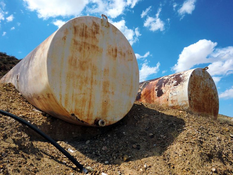 Two large rust-stained white storage tanks on gravel under a blue sky with clouds