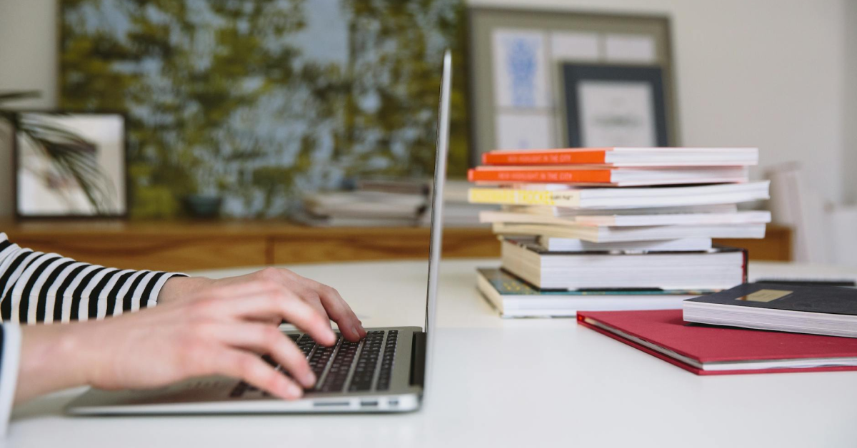 A person is typing on a laptop next to a stack of books.