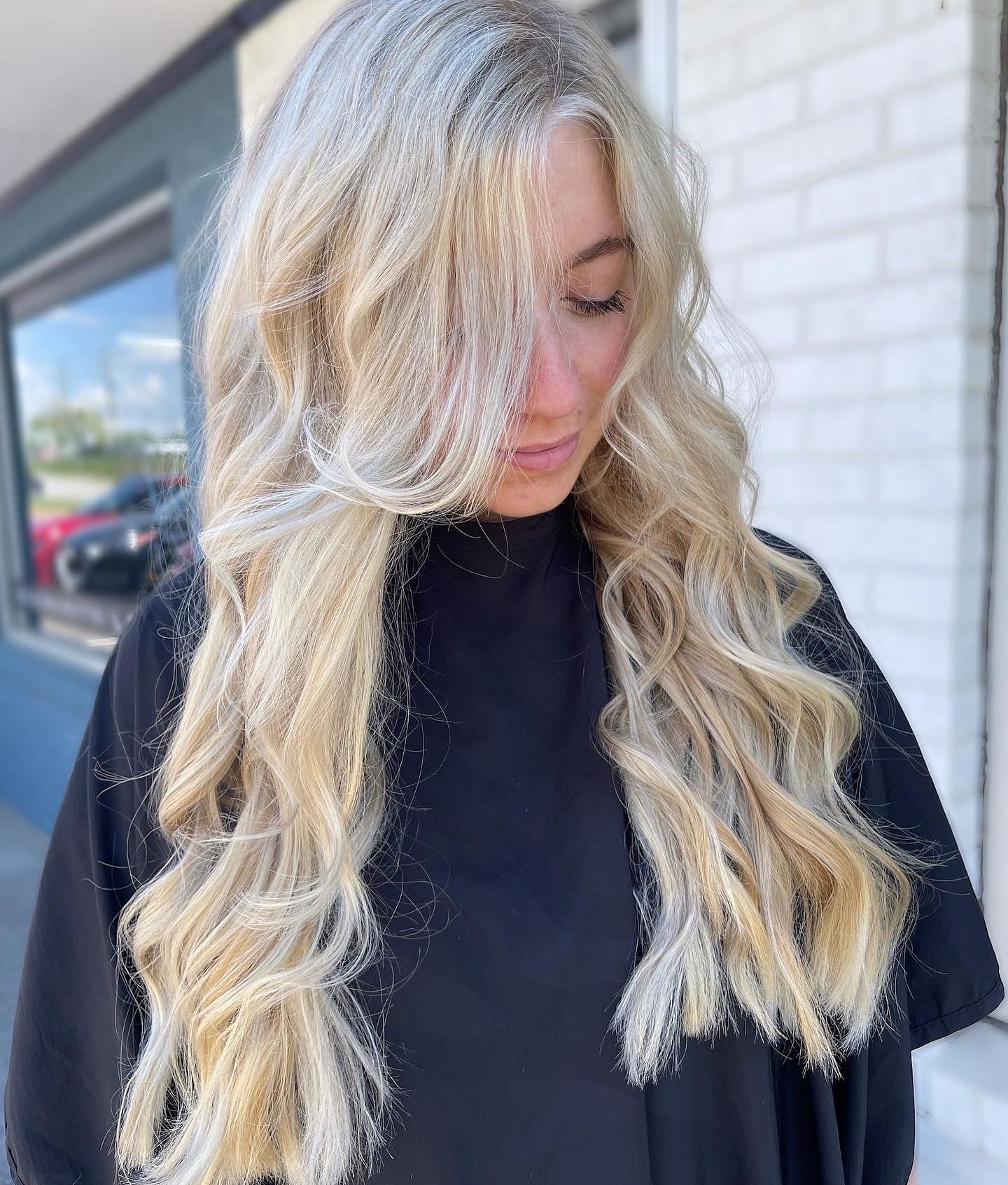 Blonde wavy hair styled on a person in a black salon cape, natural outdoor light.