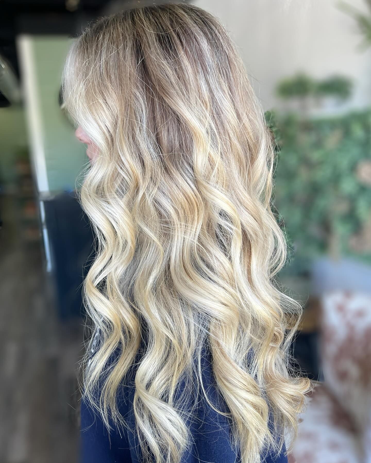 Blonde wavy hair, balayage highlights, woman's shoulder and back facing camera in salon.