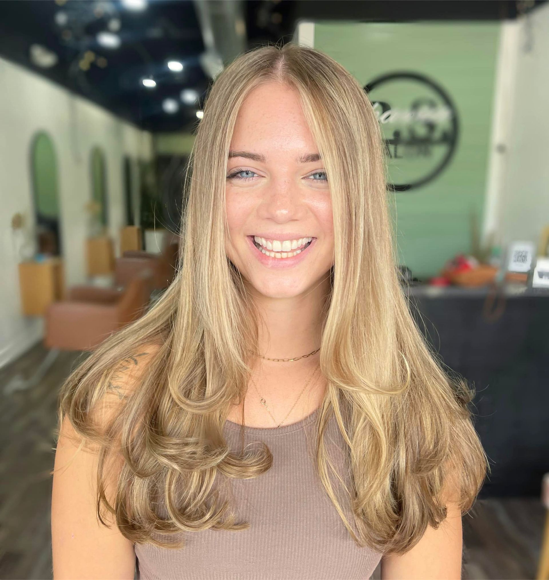 Blonde woman smiling in a hair salon. Long layered hair, tan top.