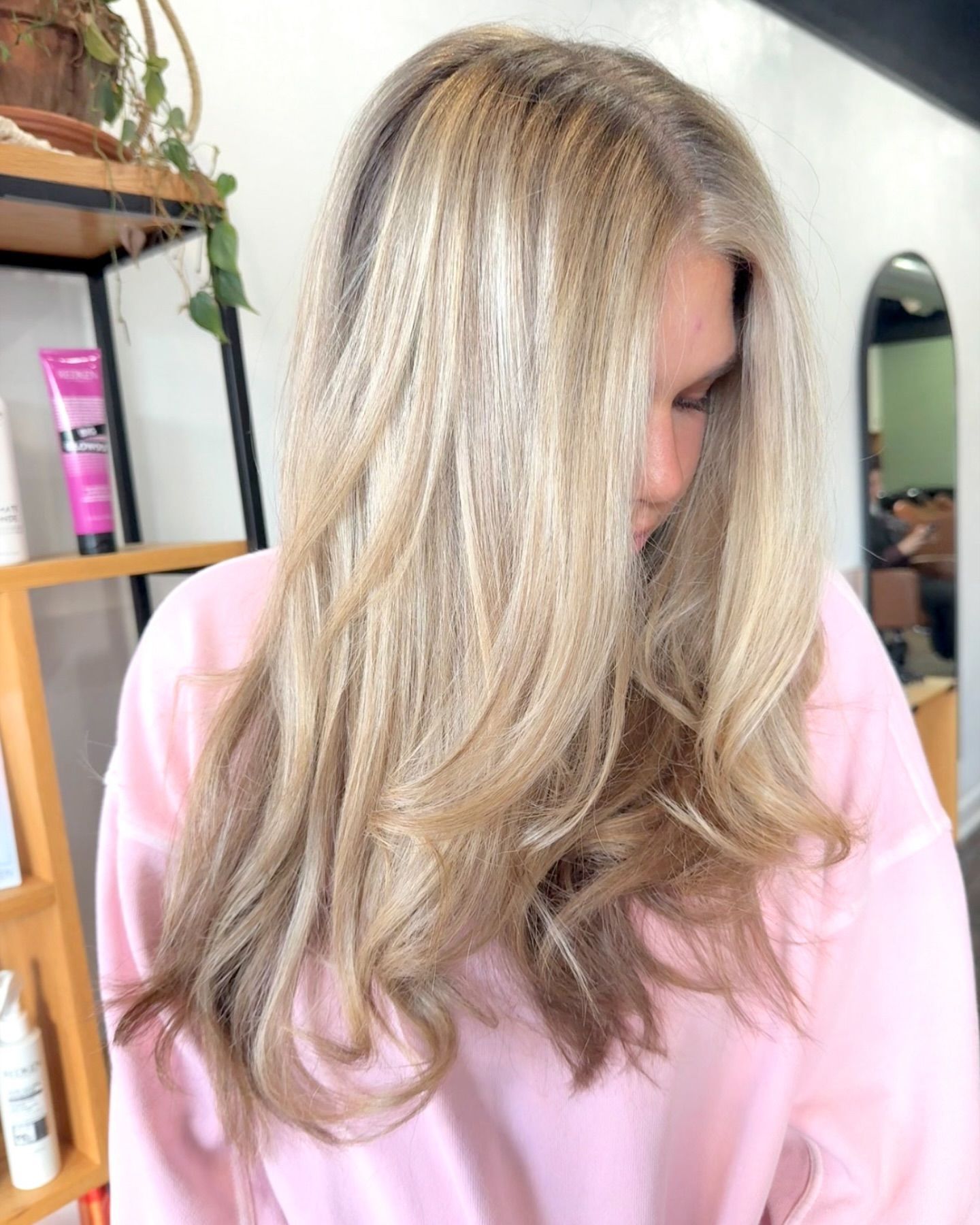 Woman with long blonde wavy hair, wearing a pink shirt, in a salon.