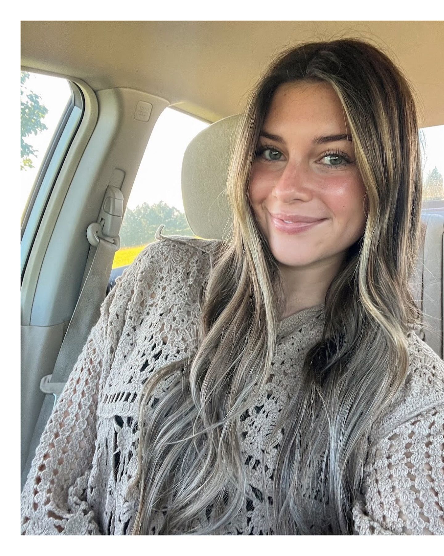 Woman smiling, sitting inside a car with sunlight streaming in. Wearing a tan, open-weave sweater.