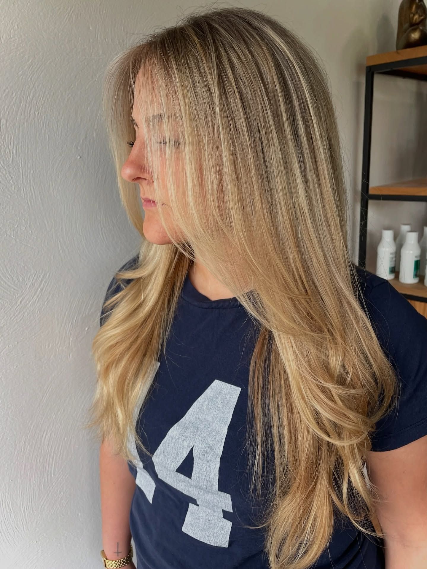 Woman with long blonde hair, wearing a navy t-shirt. She is standing next to a white wall.