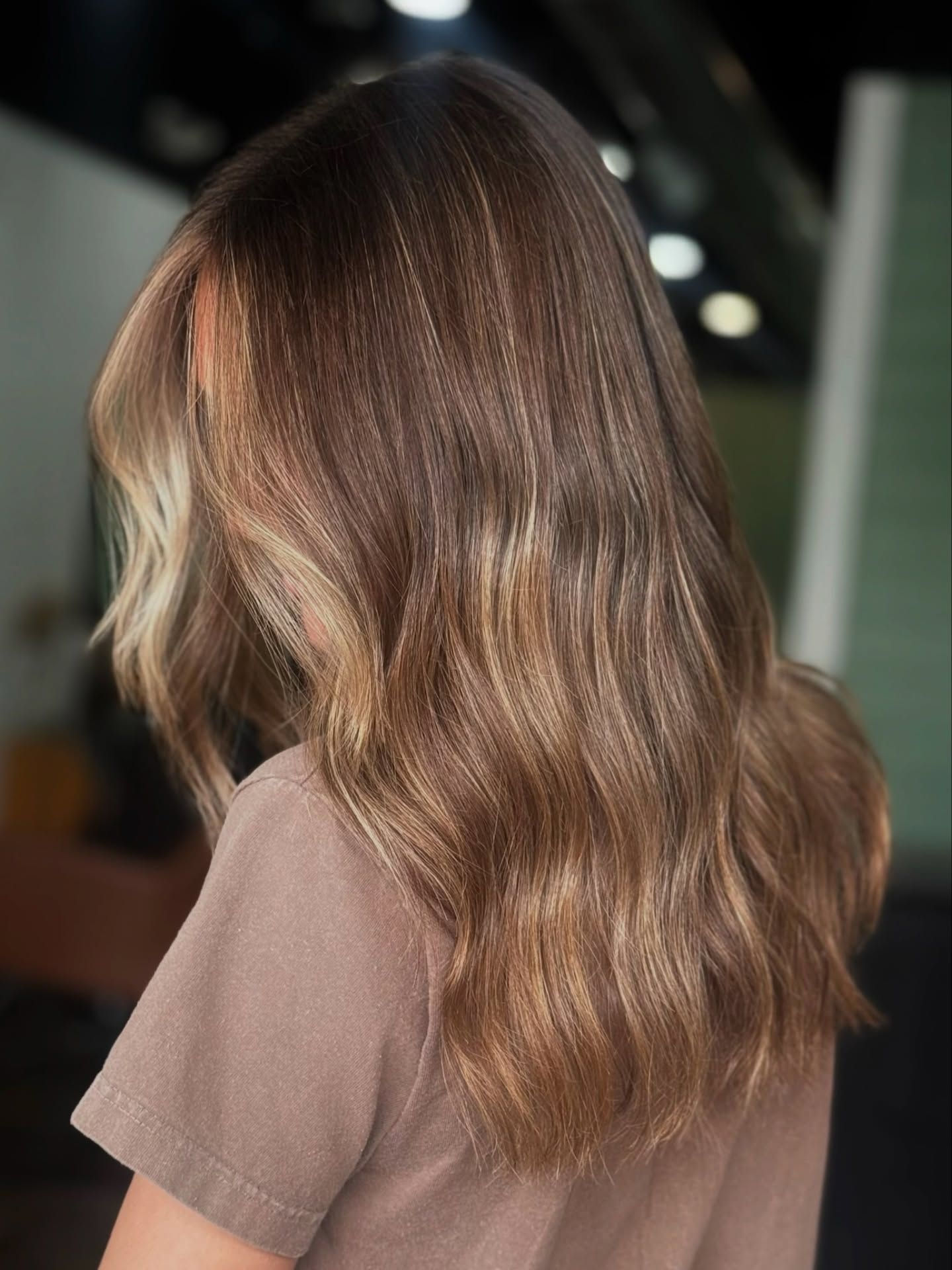 Woman with long wavy brown hair, sun-kissed highlights, and a beige top.