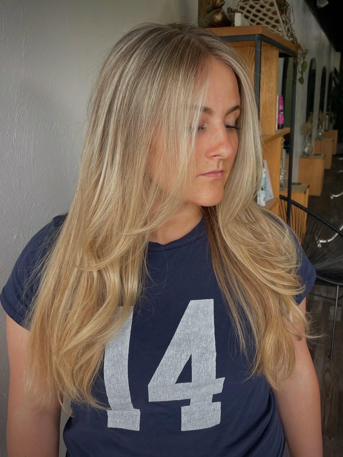 Blonde woman with long layered hair, wearing a navy t-shirt. She is in a salon setting.