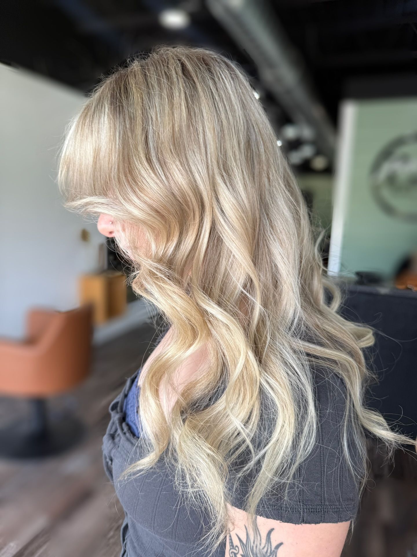 Woman with long wavy blonde hair, wearing a gray shirt, in a salon.