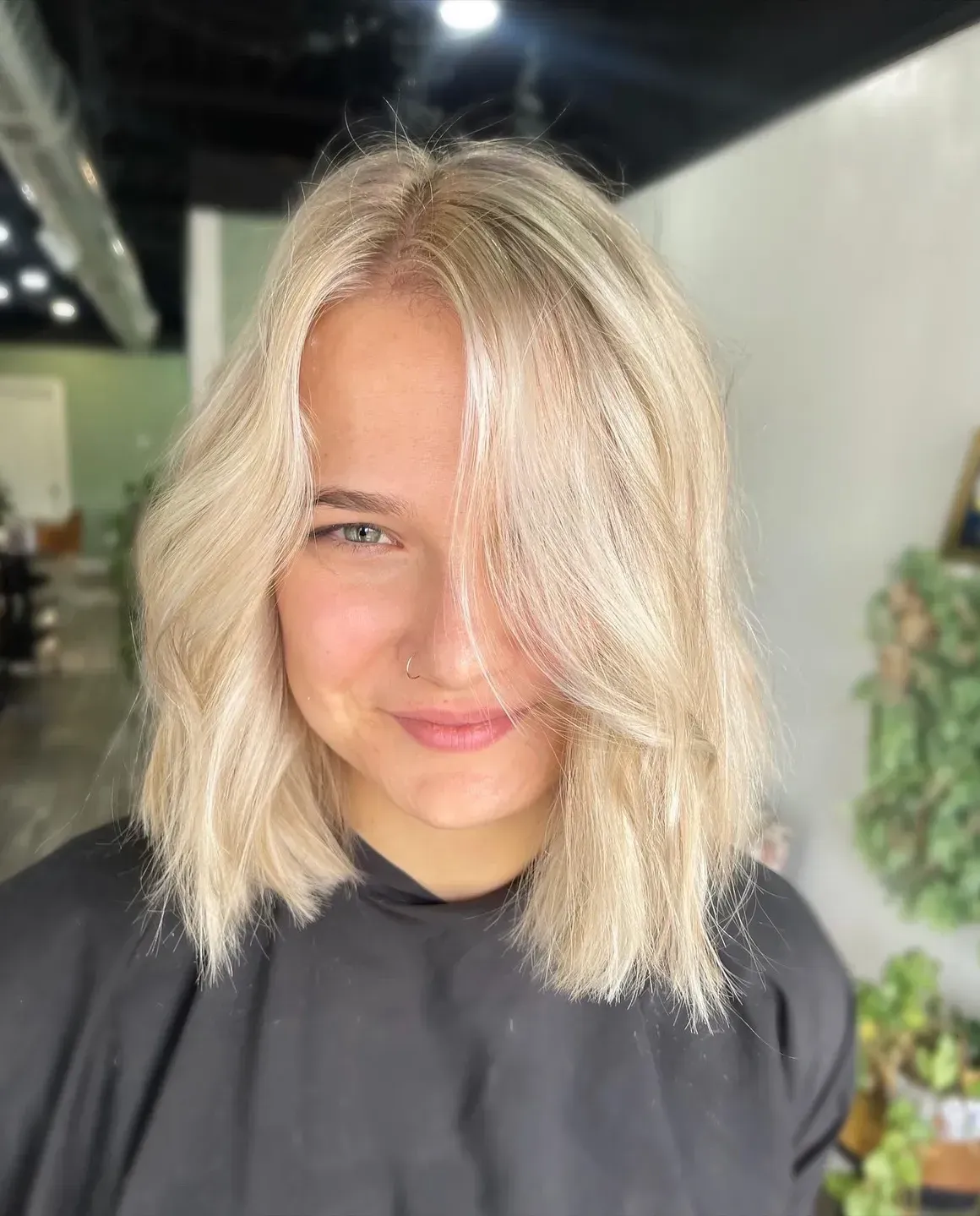 Woman with wavy, blonde shoulder-length hair smiles, nose ring, in a salon setting.