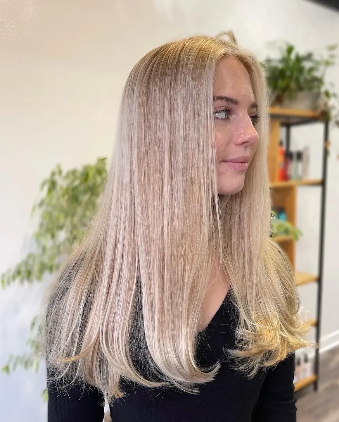 Woman with long, blonde, straight hair; black top; indoors, with plants and shelves in the background.