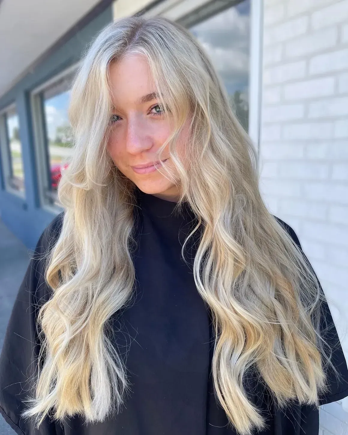 Woman with long, wavy blonde hair smiles, wearing black clothing, standing outside a building.