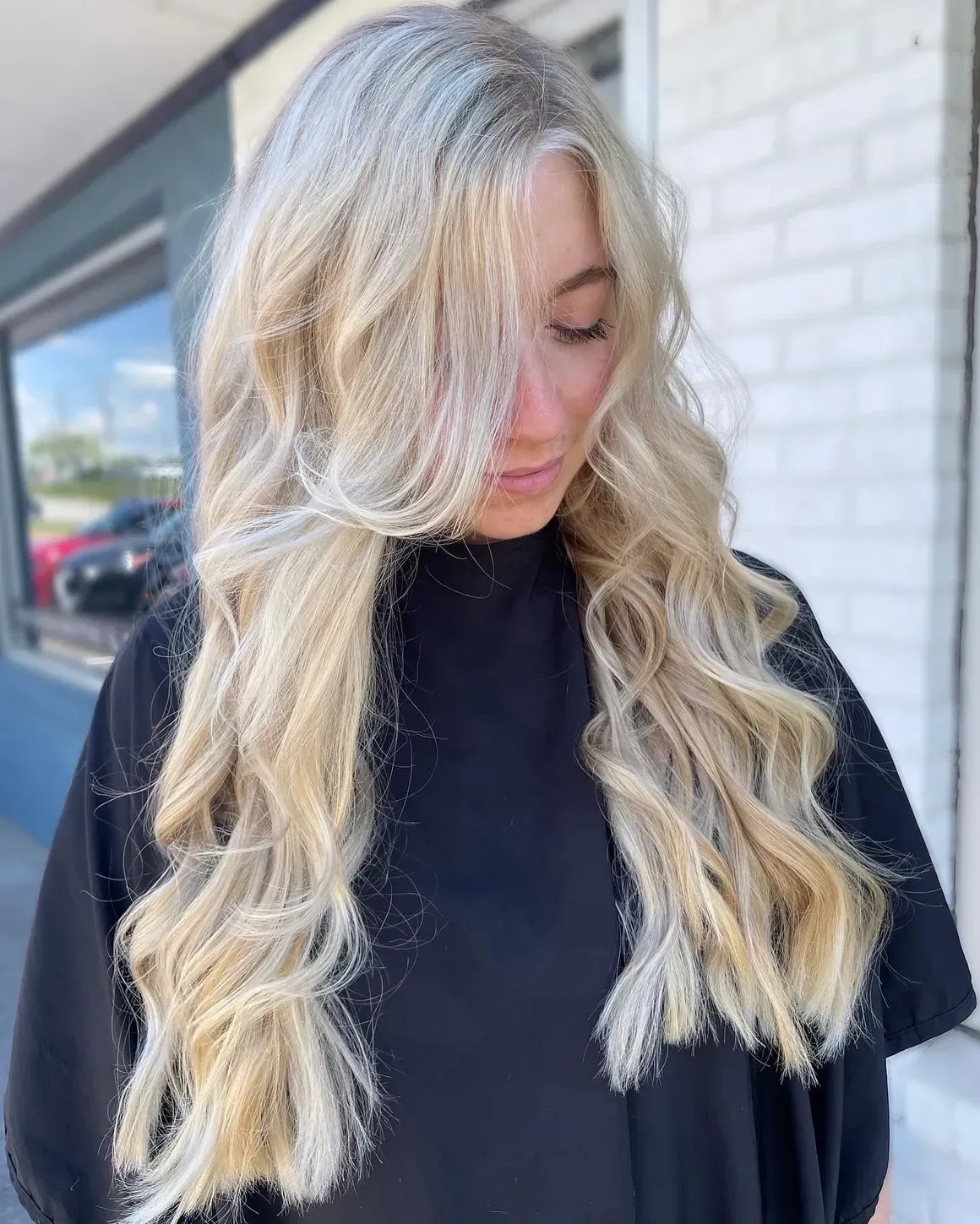 Woman with long, wavy blonde hair in front of a building; wearing a black top.