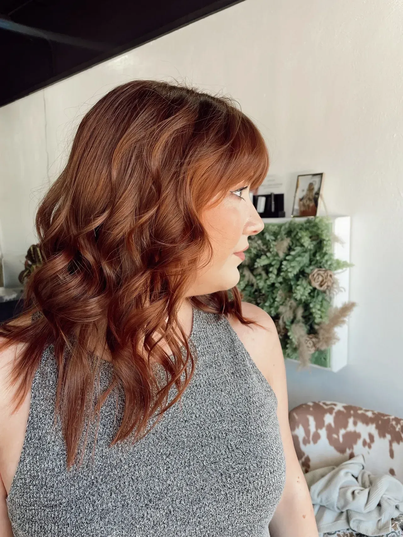 Woman with wavy auburn hair looks to the side in a gray sleeveless top. Interior with decor visible.
