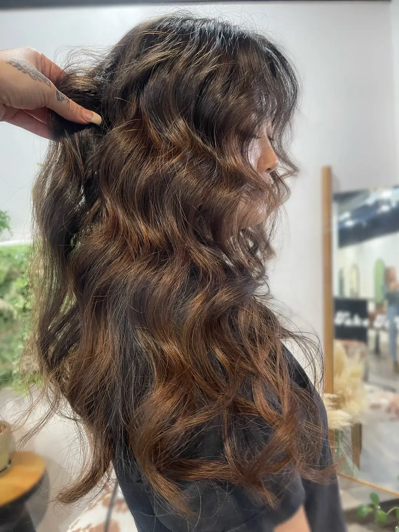 Woman with dark brown wavy hair, hand holding hair, lit salon interior.