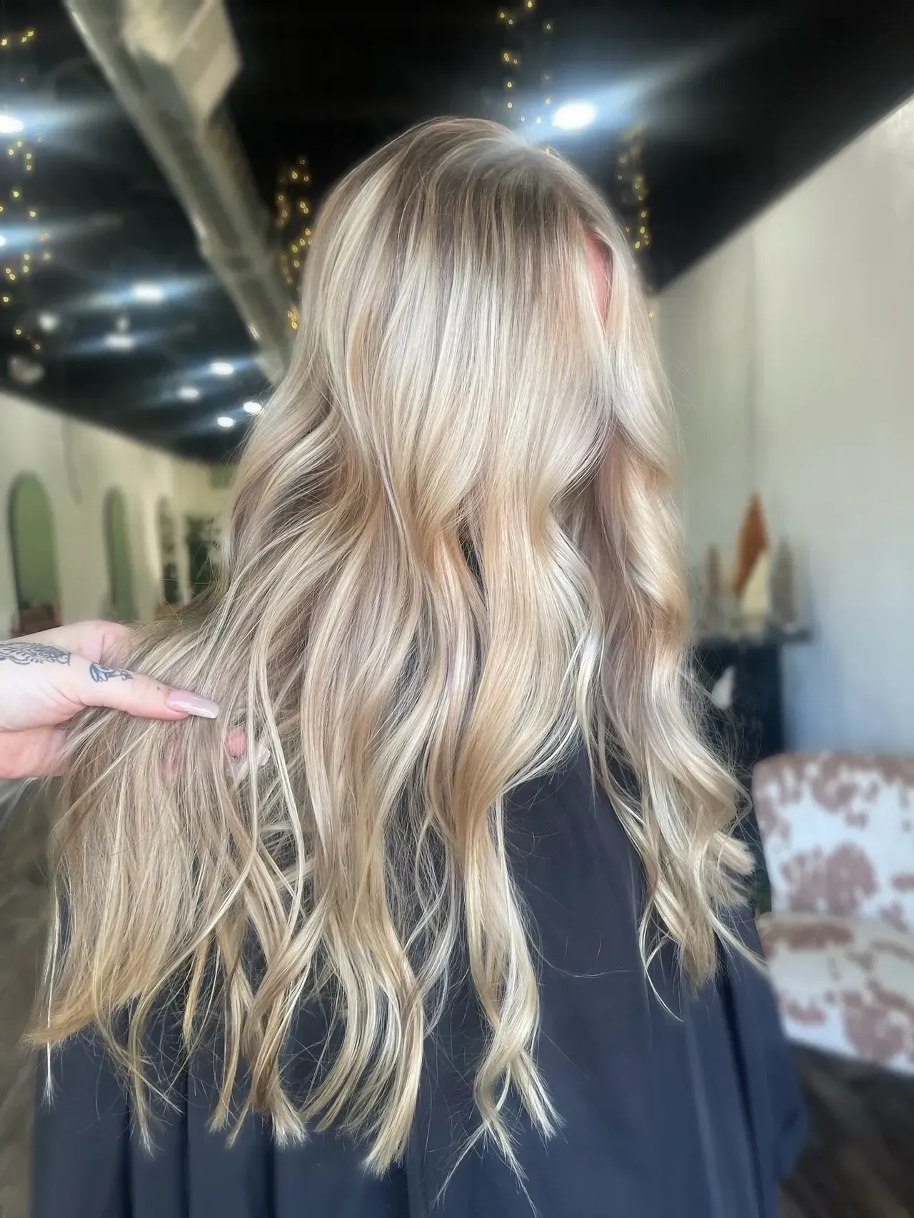 Woman with wavy blonde hair, hand touching hair. Salon interior with mirrors and cowhide chair in background.