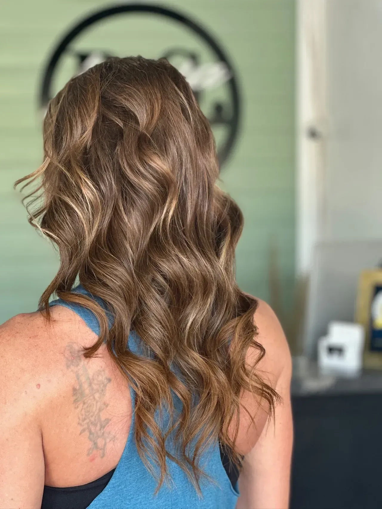 Woman with wavy, brown hair, wearing a blue tank top, standing in a salon.