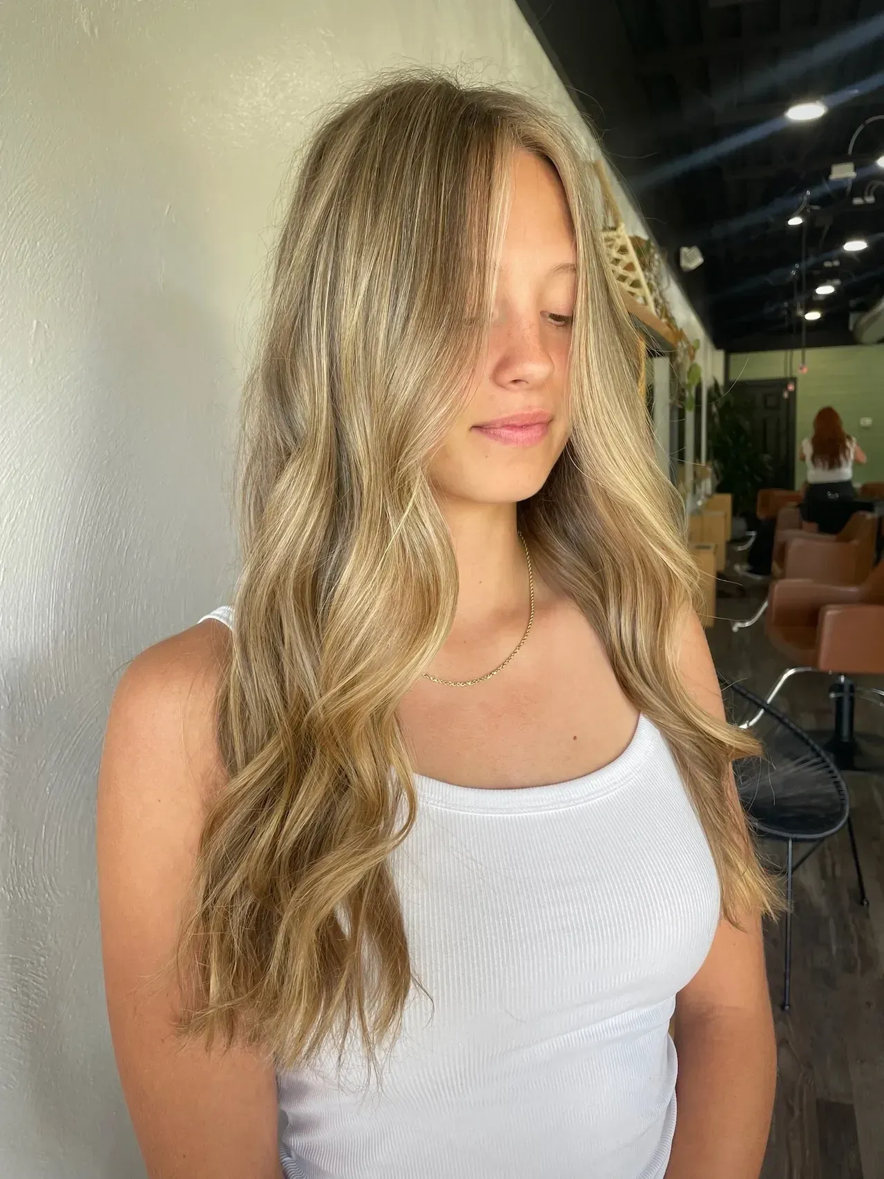 Woman with wavy, blonde hair, wearing a white tank top, in a salon.