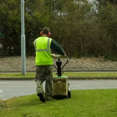A gardener mowing a roadside grass verge in Halfwaybush, Dunedin, to maintain a tidy street-front area.
