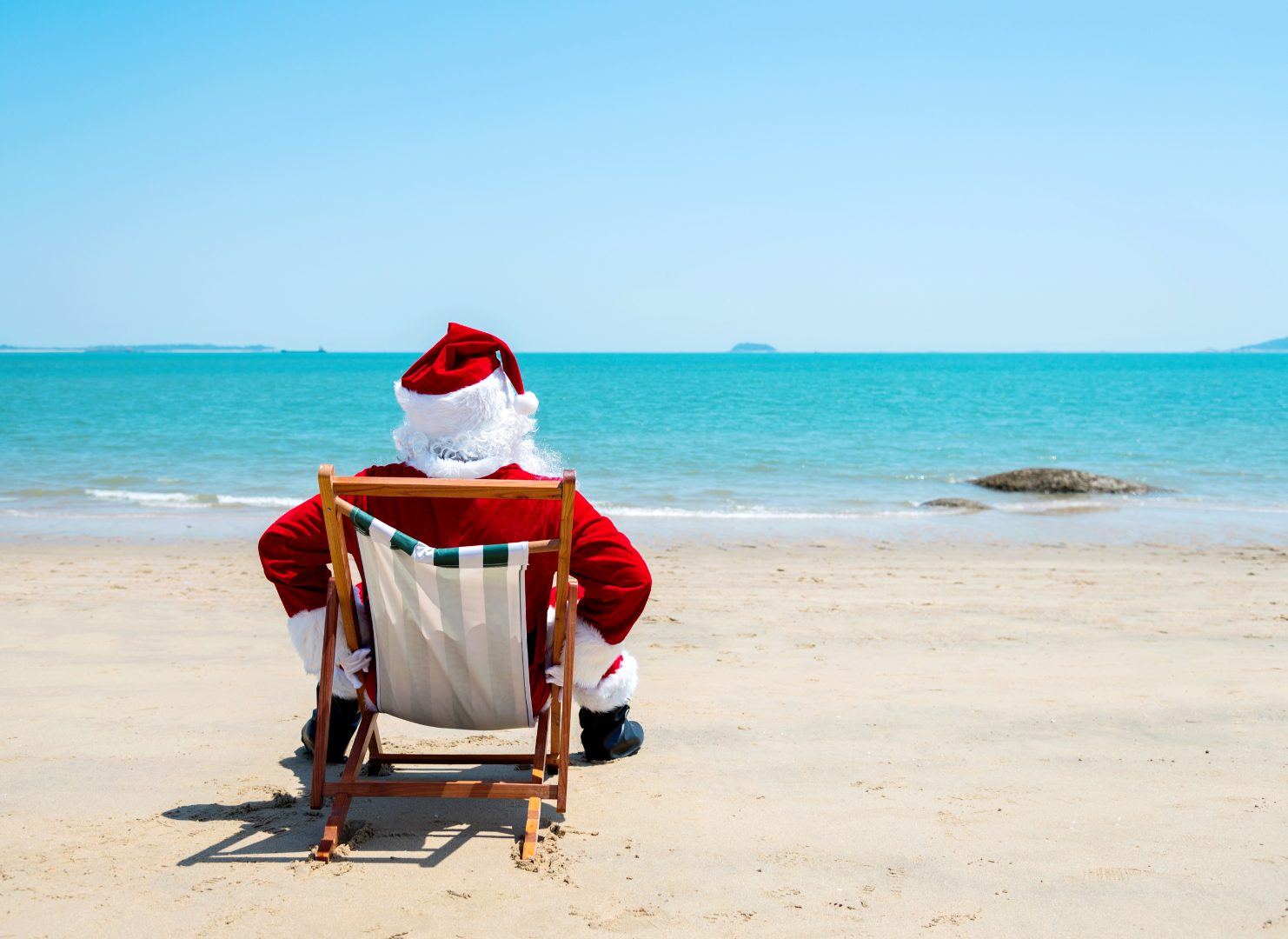 Papai Noel está sentado em uma cadeira na praia olhando para o oceano.-Hotel-Praia-Dourada-Maragogi-Park