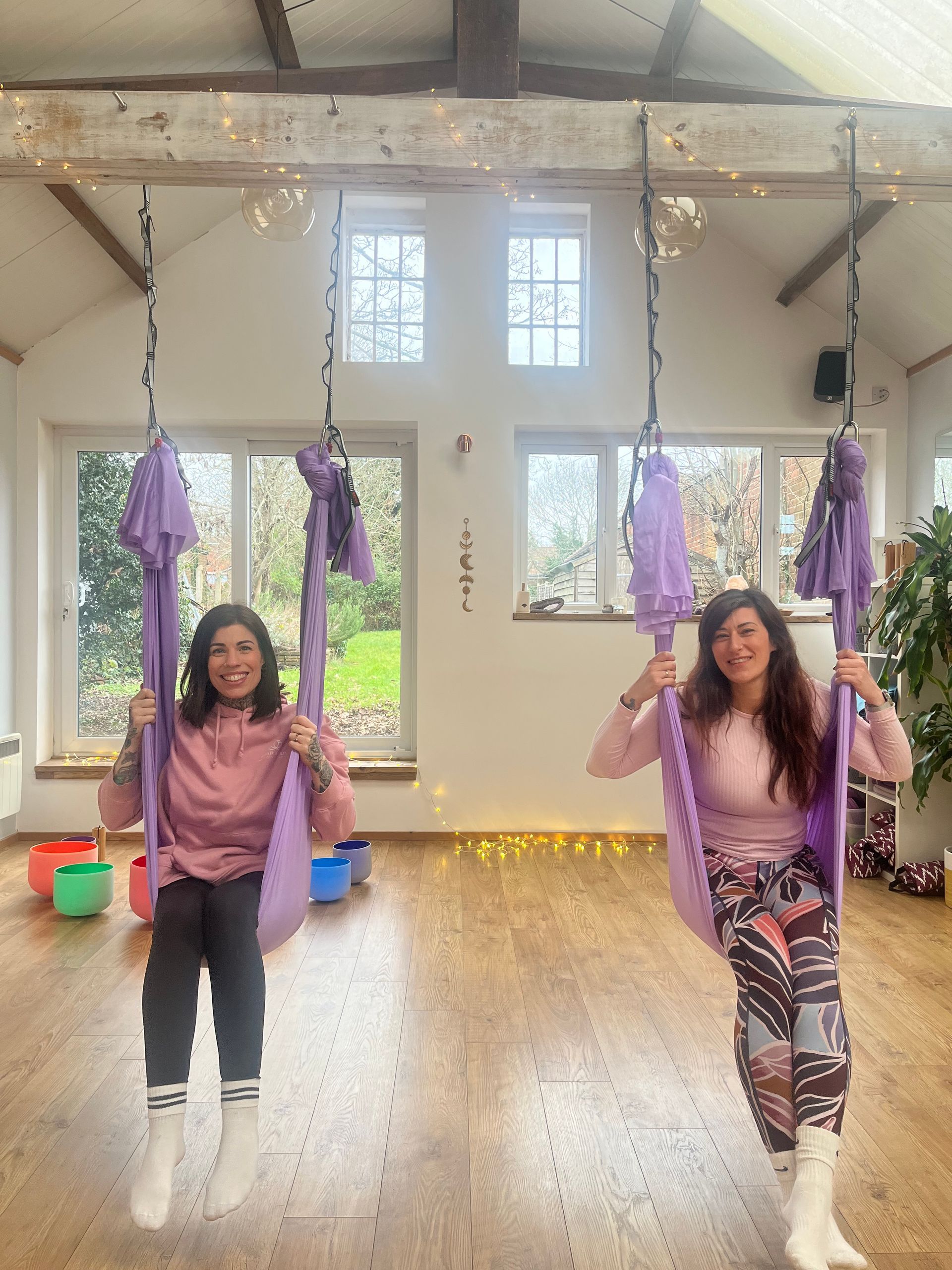 Two women smiling, seated in purple aerial yoga swings in a bright room with windows and lights.
