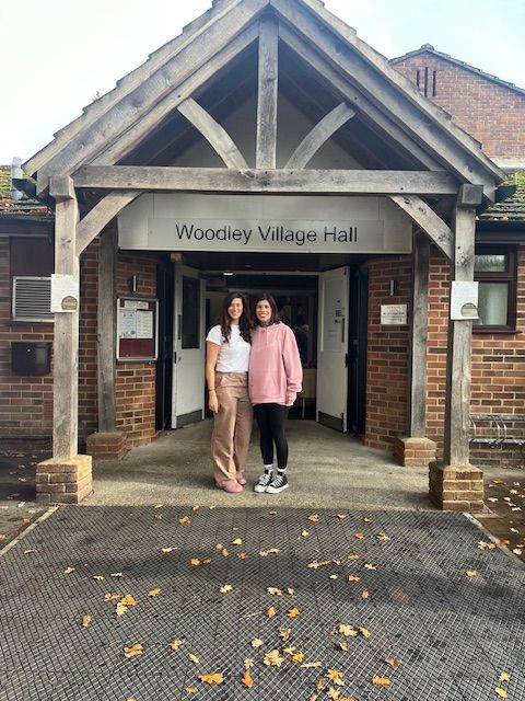 Two people stand in front of Woodley Village Hall entrance, beneath a wooden porch.