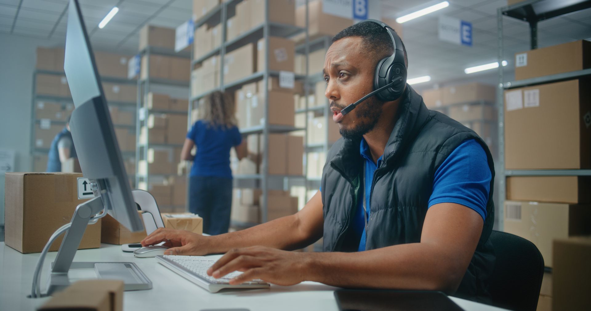 A warehouse worker wearing a headset types at a computer desk in a facility filled with storage shelves and cardboard boxes.