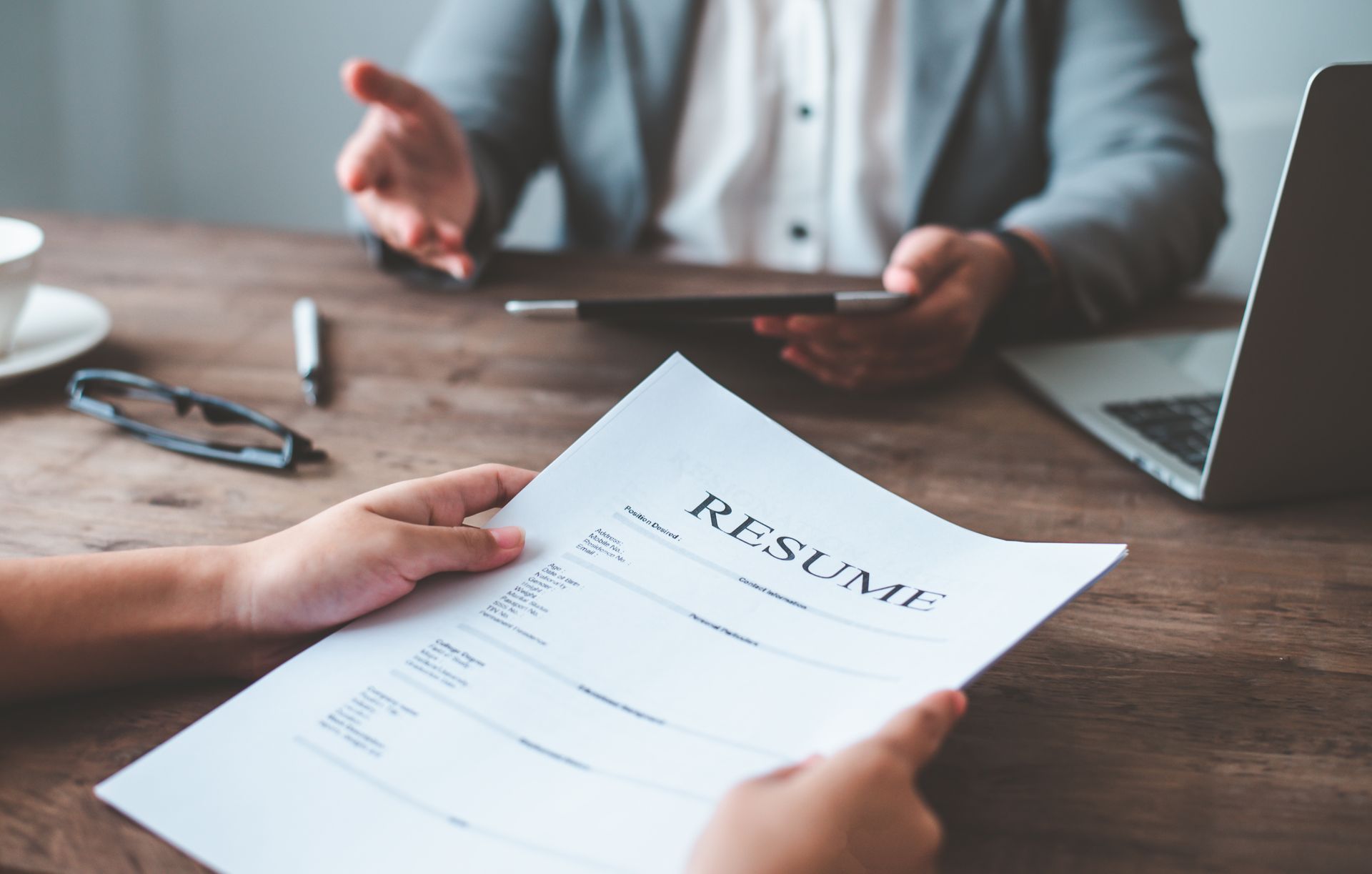 A person holds a resume across a wooden desk while an interviewer gestures during a professional meeting.