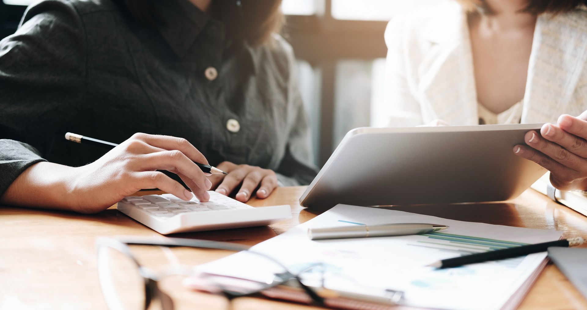 Two people working together; one uses a calculator, the other holds a tablet. Papers, glasses on the desk.