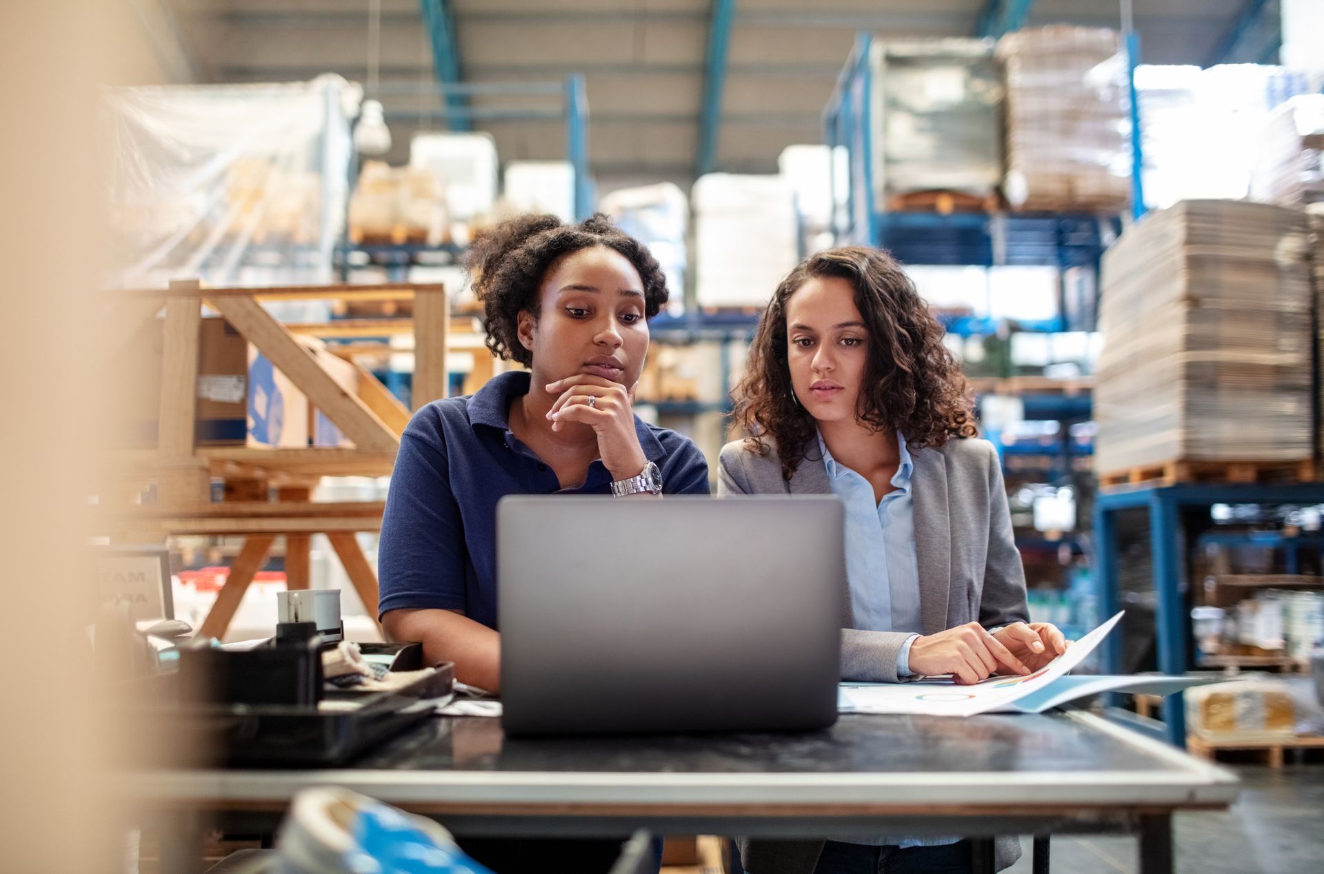 Two women are sitting at a table in a warehouse looking at a laptop computer.