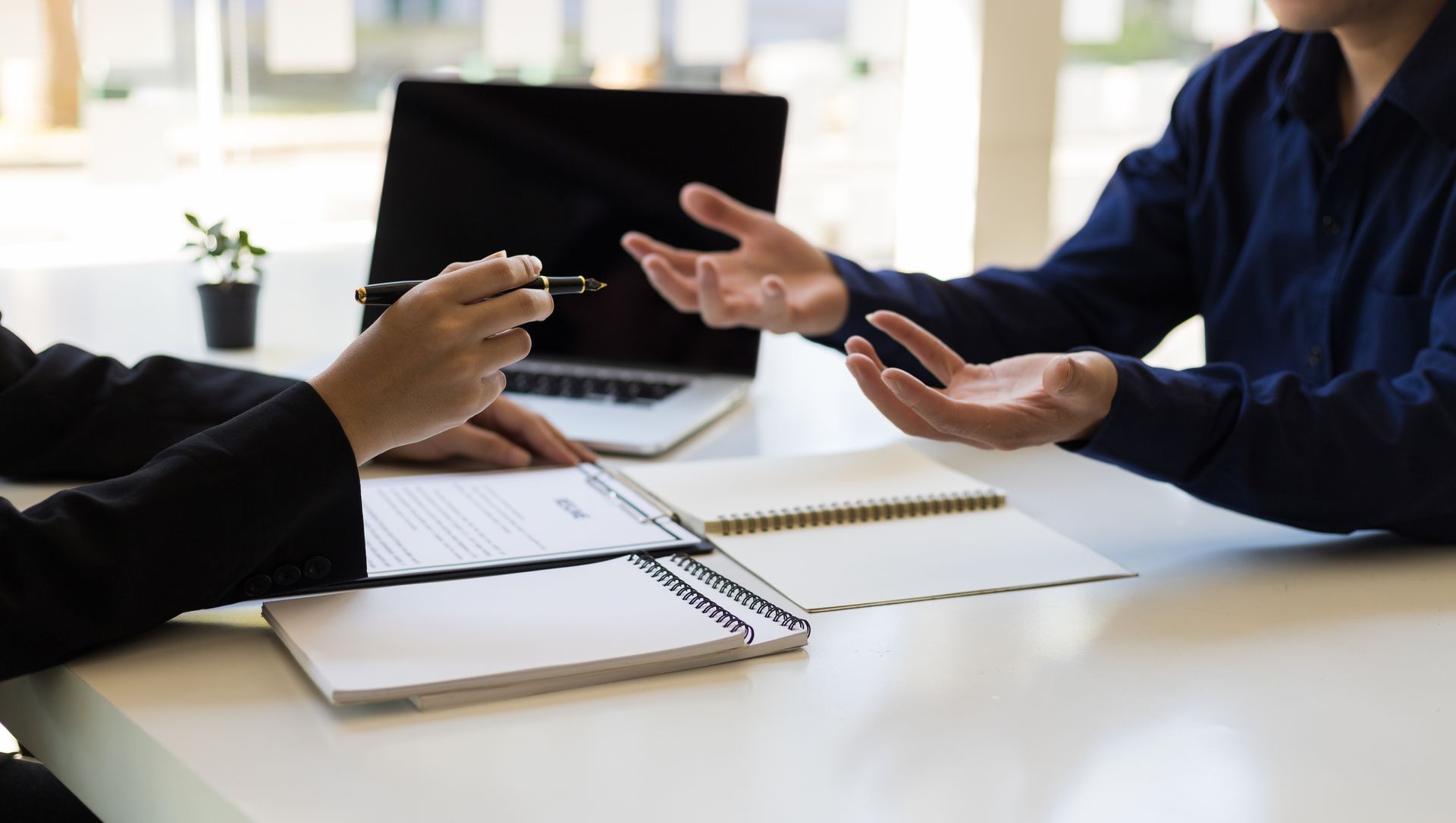 Two people at a desk, one holding a pen and papers, the other gesturing with open hands, in a professional setting.