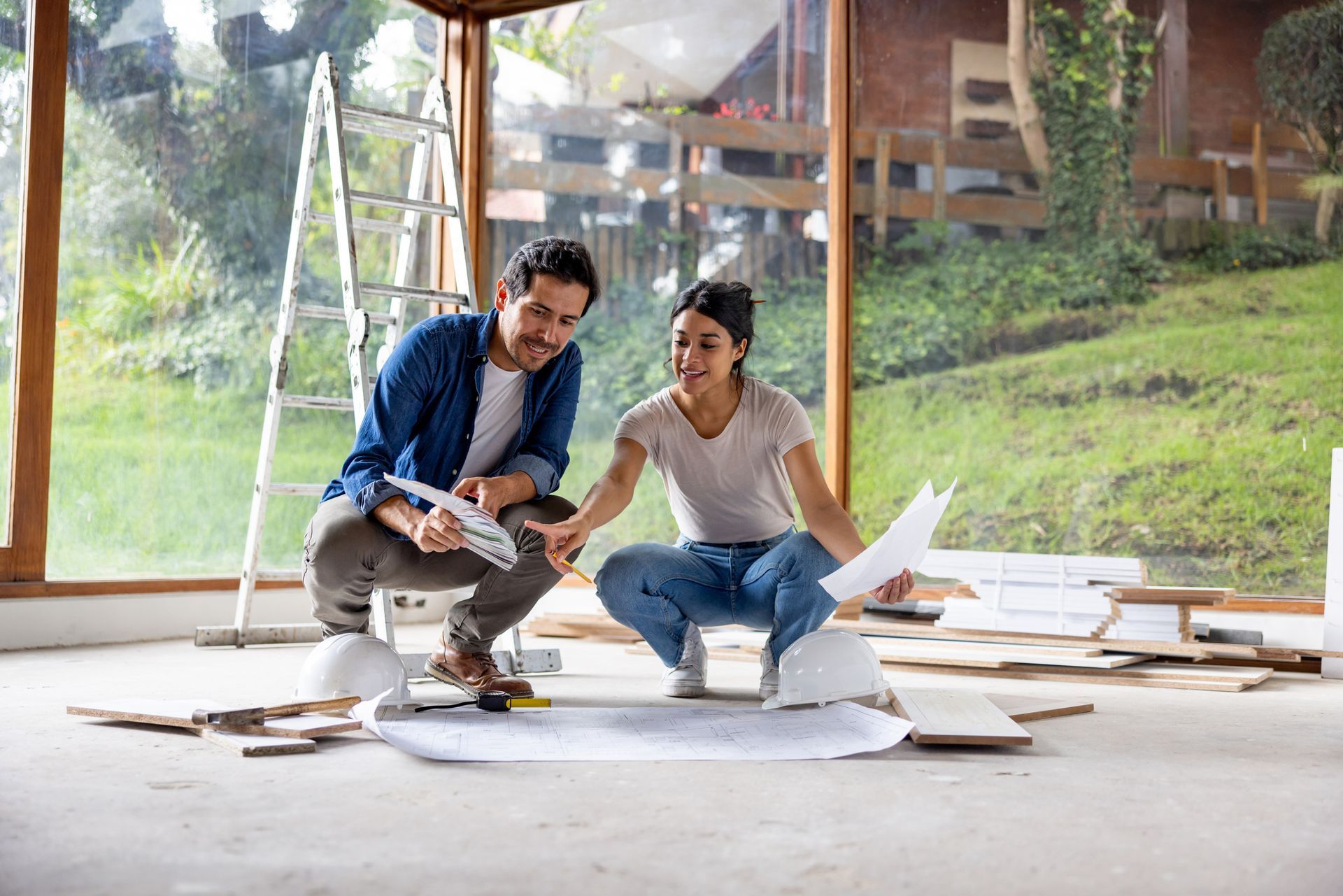 Couple reviewing blueprints in a sunlit room under construction; smiling, pointing.
