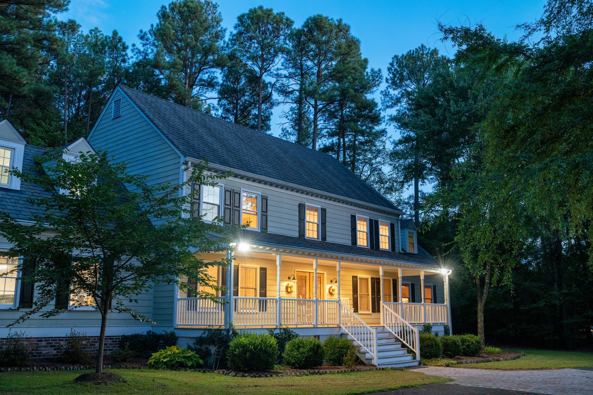 Two-story light blue house with porch lit up at dusk, surrounded by trees.