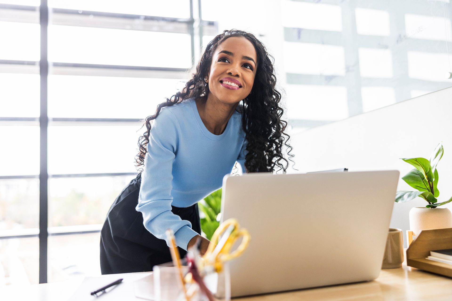 Woman smiling, leaning on desk with laptop, plant and office supplies.
