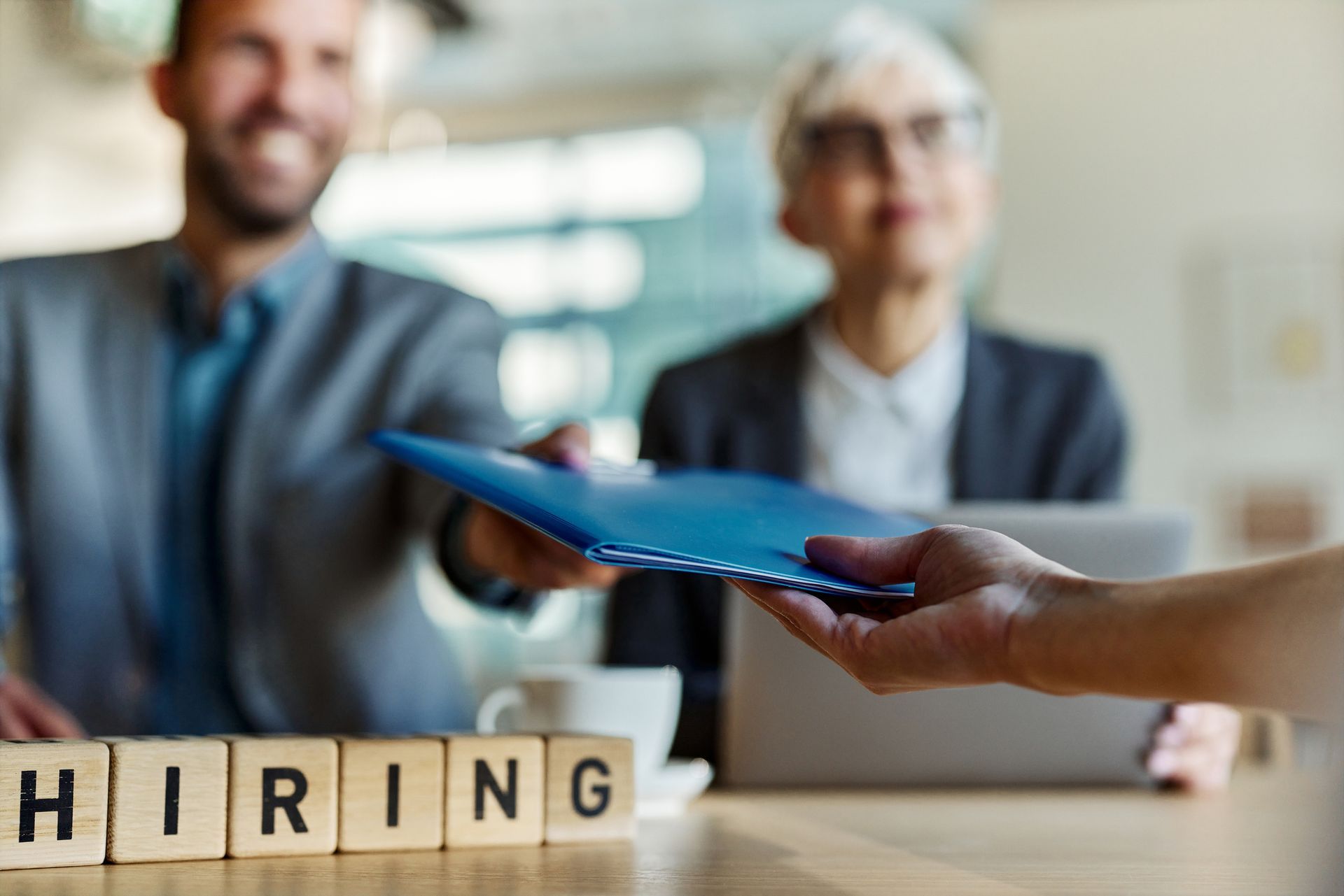 Close-up of a wooden block spelling “hiring” with an unseen candidate submitting a resume.