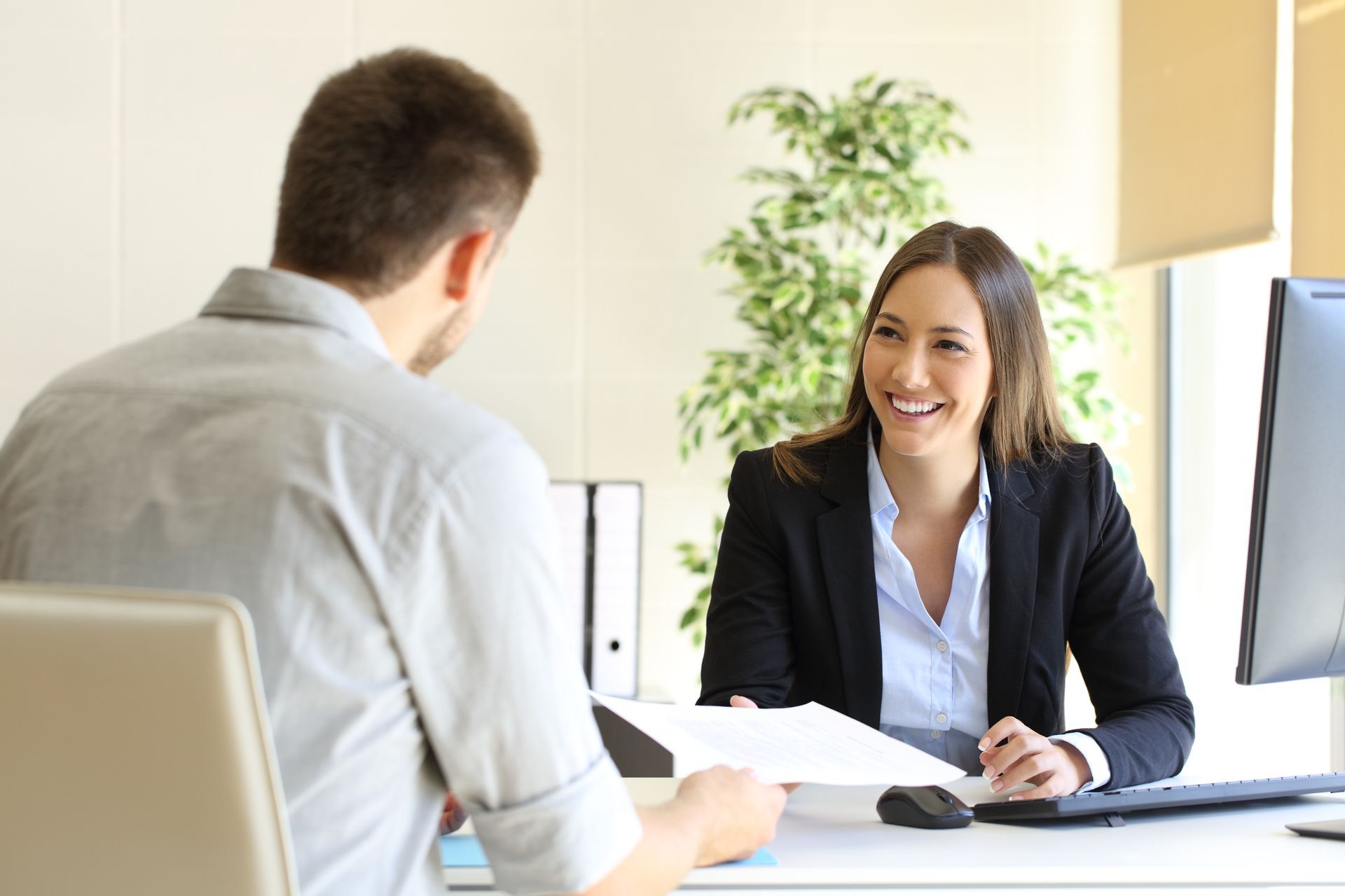Woman in a suit smiling, handing documents to a man in a light shirt across a desk. Office setting.