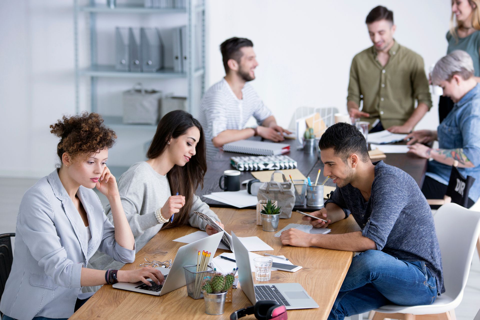 Group of diverse people at a table in an office, working on laptops and discussing.