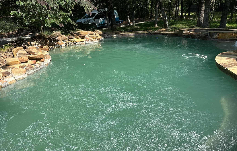 A turquoise pool with water bubbling, bordered by rock and trees.
