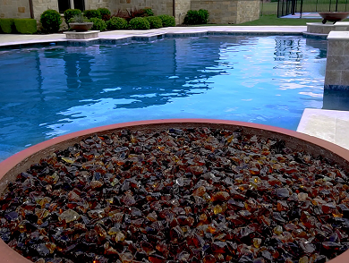 Fire pit with brown glass rocks, next to a pool with a blue surface.