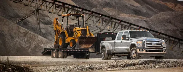 Truck towing a trailer carrying a yellow backhoe in a quarry.