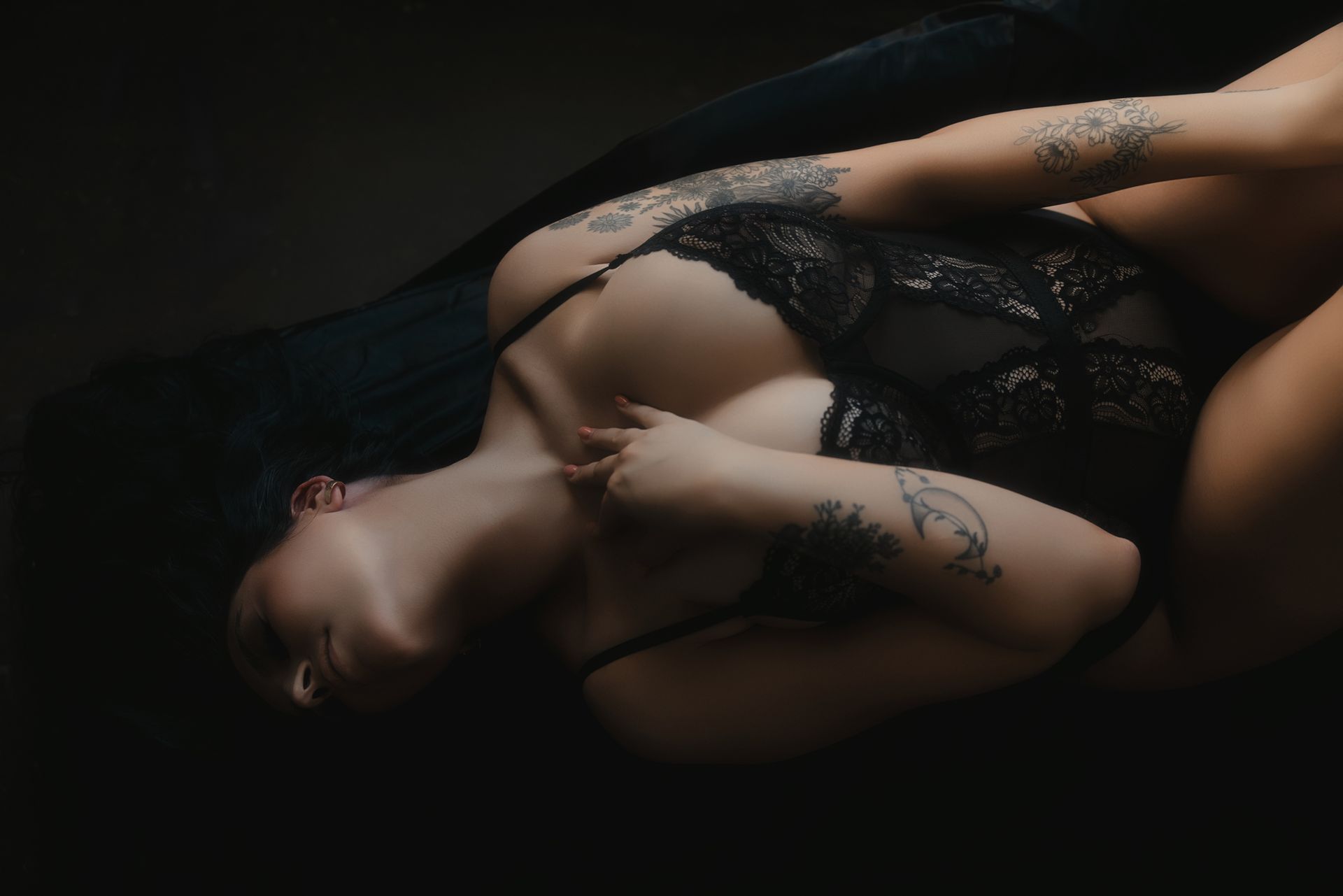 Woman in black lingerie reclines, arms crossed, looking away. Dark setting with visible tattoos.