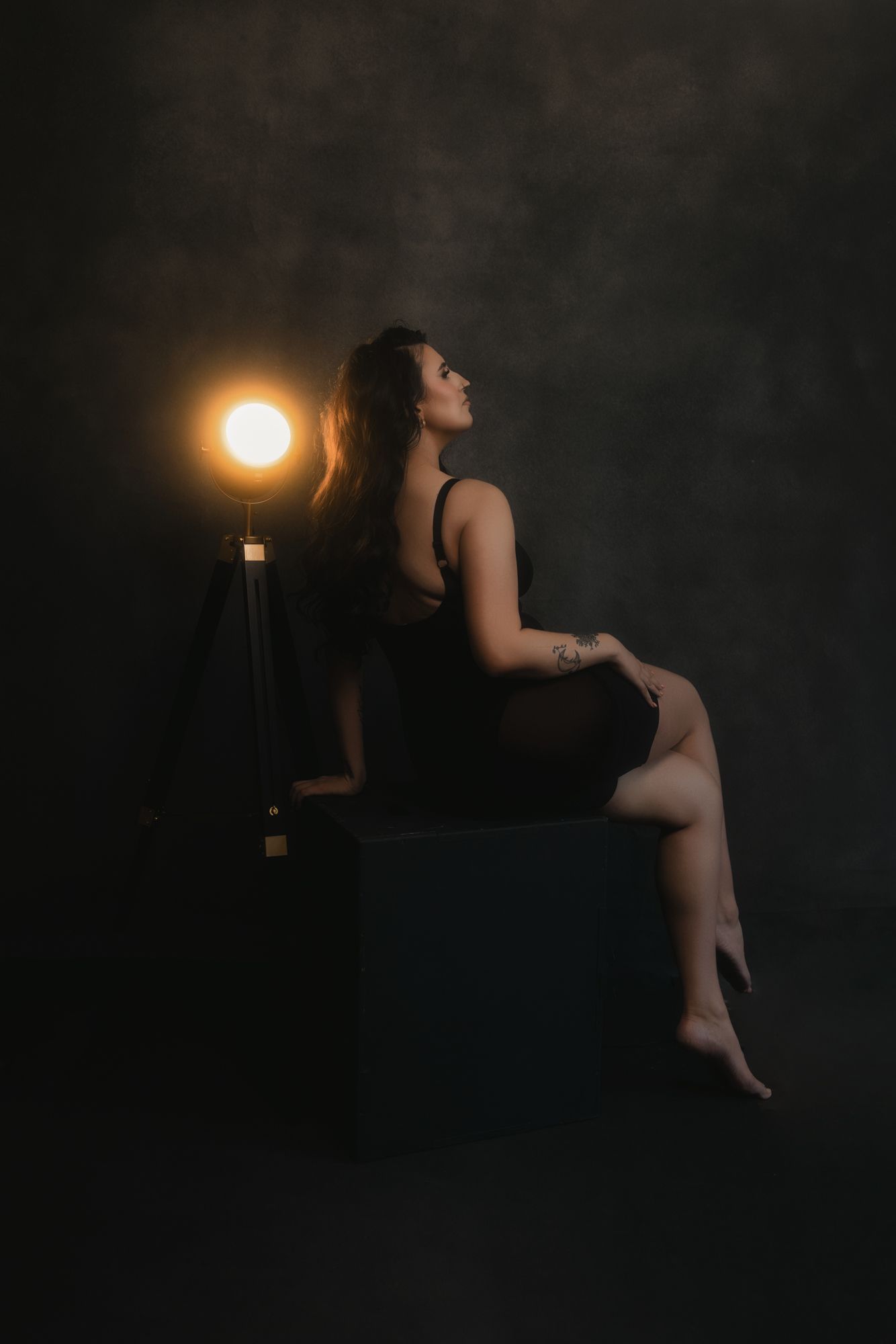 Woman in black lingerie sits on a black box, posing with a studio light. Dark background with a warm glow on the subject's side.